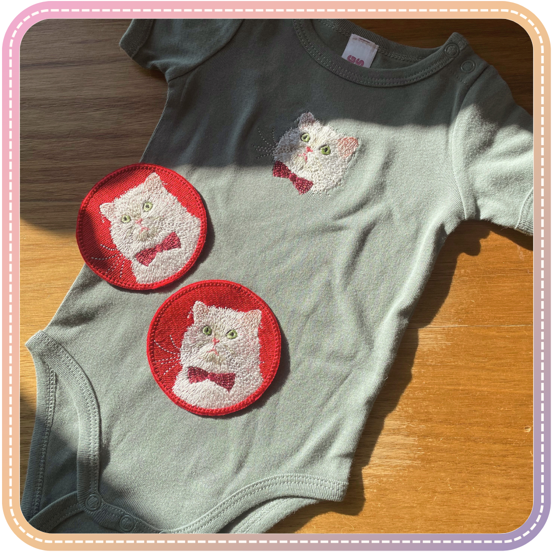 a baby onesie and two embroidered patches featuring a white cat with a red bowtie