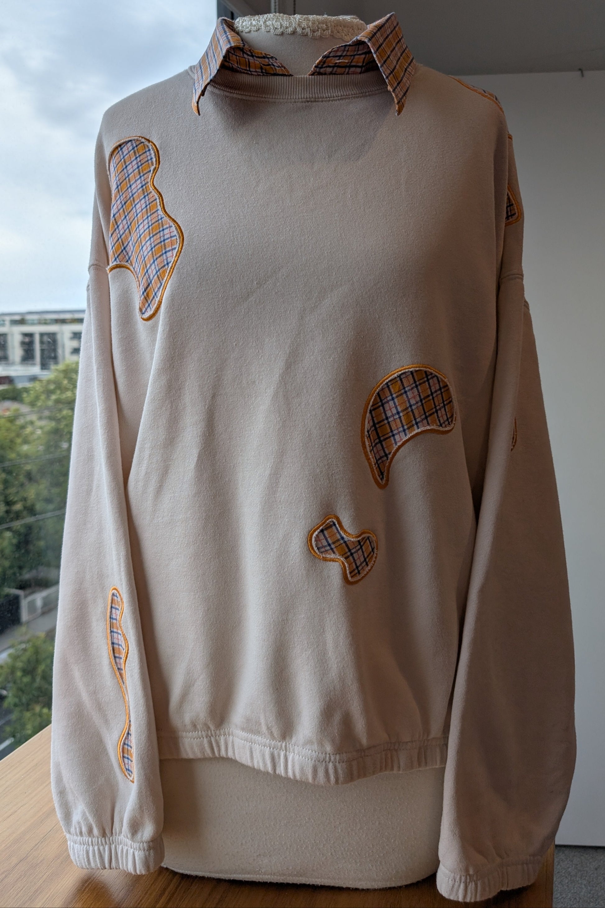beige crew neck upcycled with embroidery blob cut outs over a blue and orange checkered shirt.